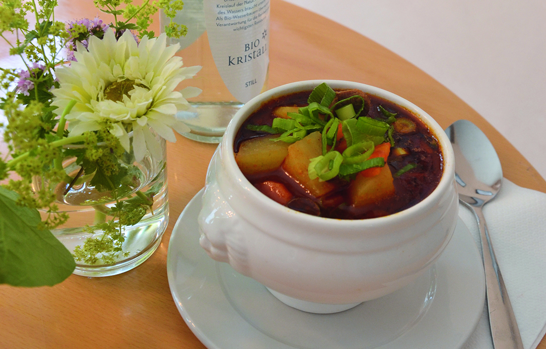 Dampfende Gemüsesuppe in einer weißen Suppentasse, serviert auf einem Holztisch mit Blumenarrangement und Besteck im Staufen Suppentöpfle in Göppingen.