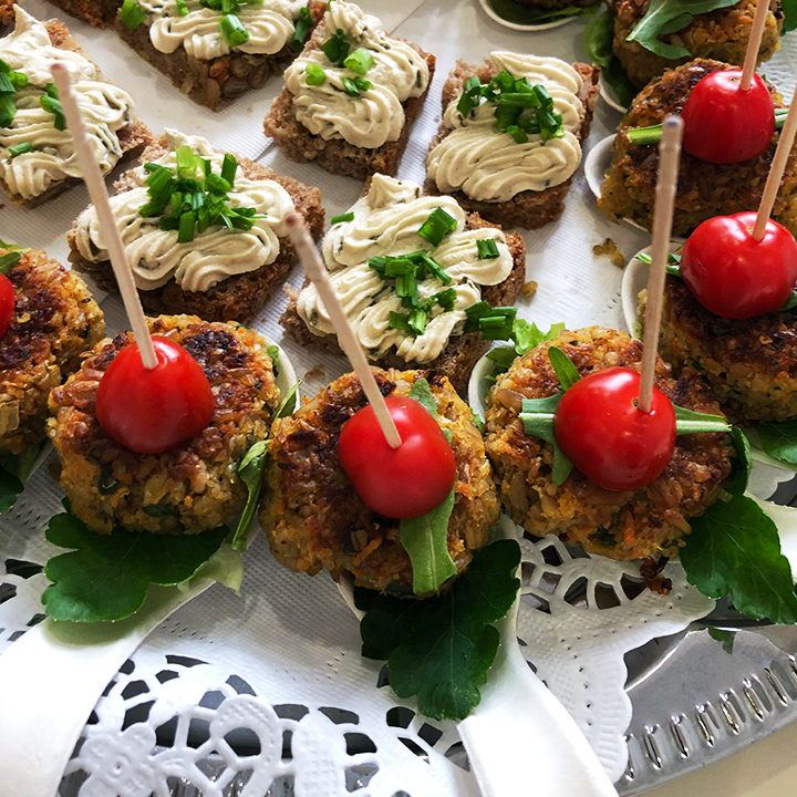 Platte mit herzhaften Häppchen: kleine Brotstücke mit Creme und Schnittlauch sowie vegetarisch wirkende Mini‑Frikadellen mit Kirschtomaten-Spieß auf Salatblättern.